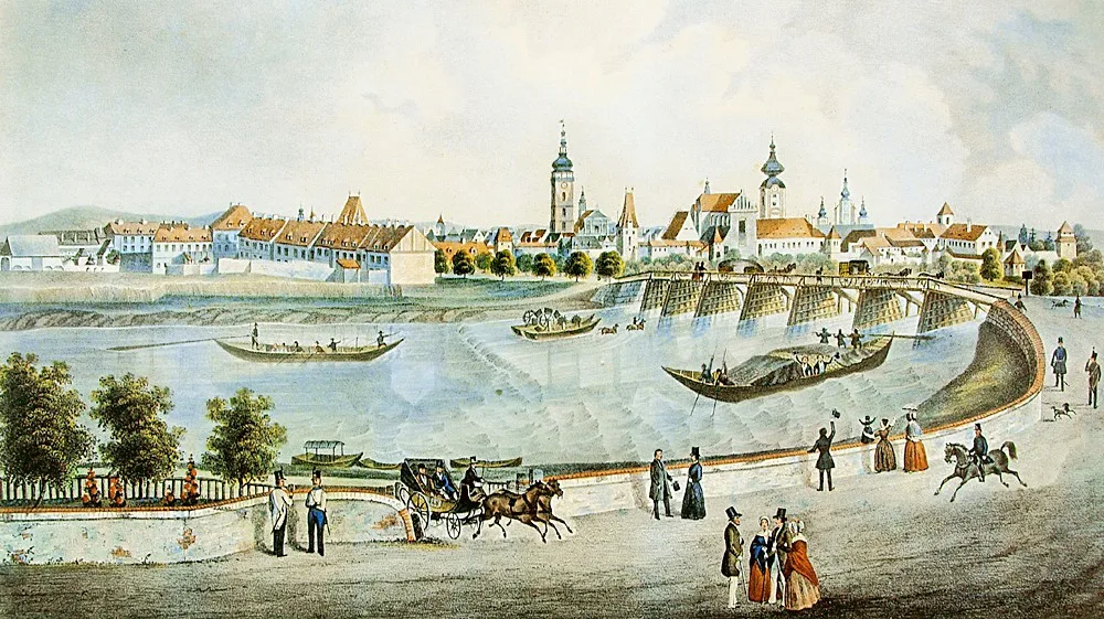 Veduta města od západu z poloviny 19. století. V popředí Vltava a lodě se starým dřevěným Dlouhým mostem. Romantické vltavské nábřeží možná tak romantické bývalo