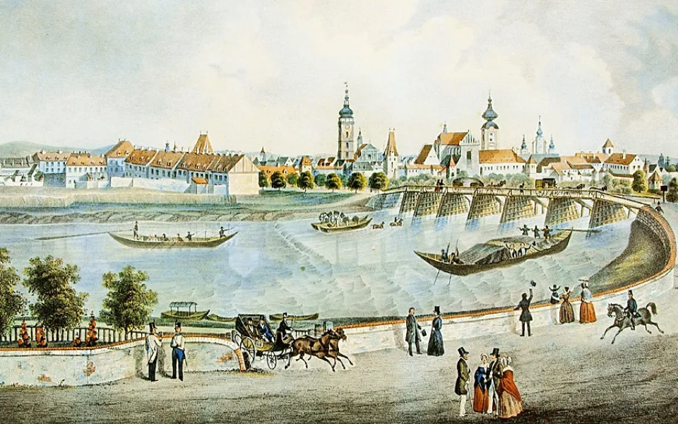 Veduta města od západu z poloviny 19. století. V popředí Vltava a lodě se starým dřevěným Dlouhým mostem. Romantické vltavské nábřeží možná tak romantické bývalo